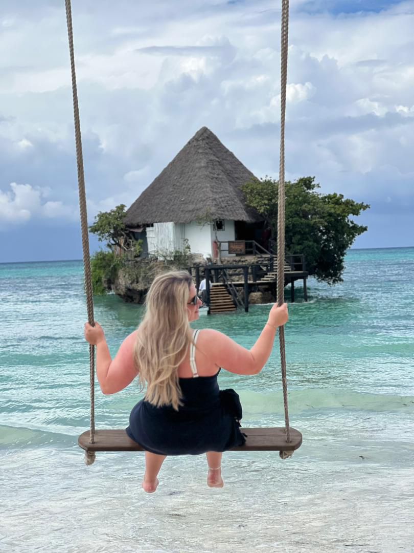 A traveler on a beach swing facing a traditional thatched Zanzibar sea pavilion